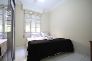 Ein Schlafzimmer mit einem Bett, zwei Fenstern mit Jalousien und einem Fliesenboden.