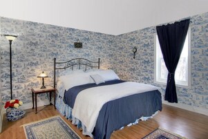 Un dormitorio con cama, mesita de noche, lámpara, ventana con cortinas y papel tapiz estampado.