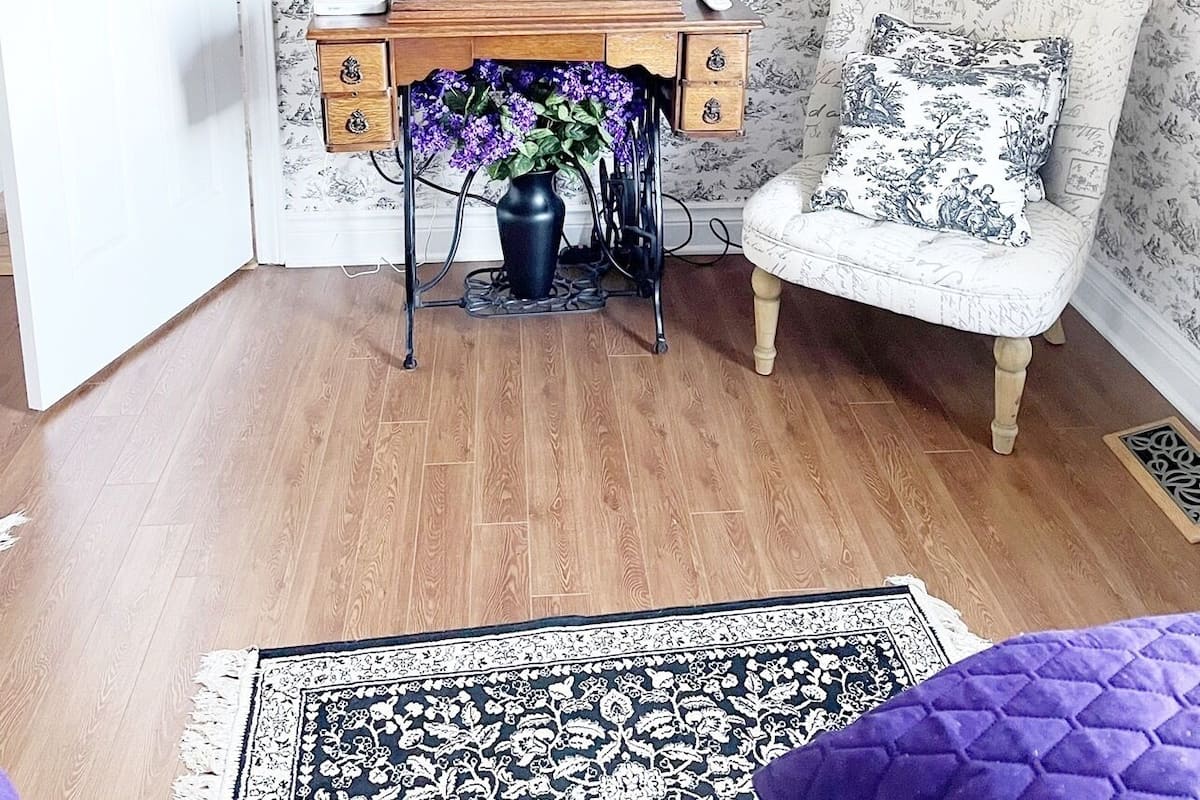 A room with a wooden desk, a flat-screen TV, a chair with a patterned cushion, a vase with flowers, and a patterned rug.