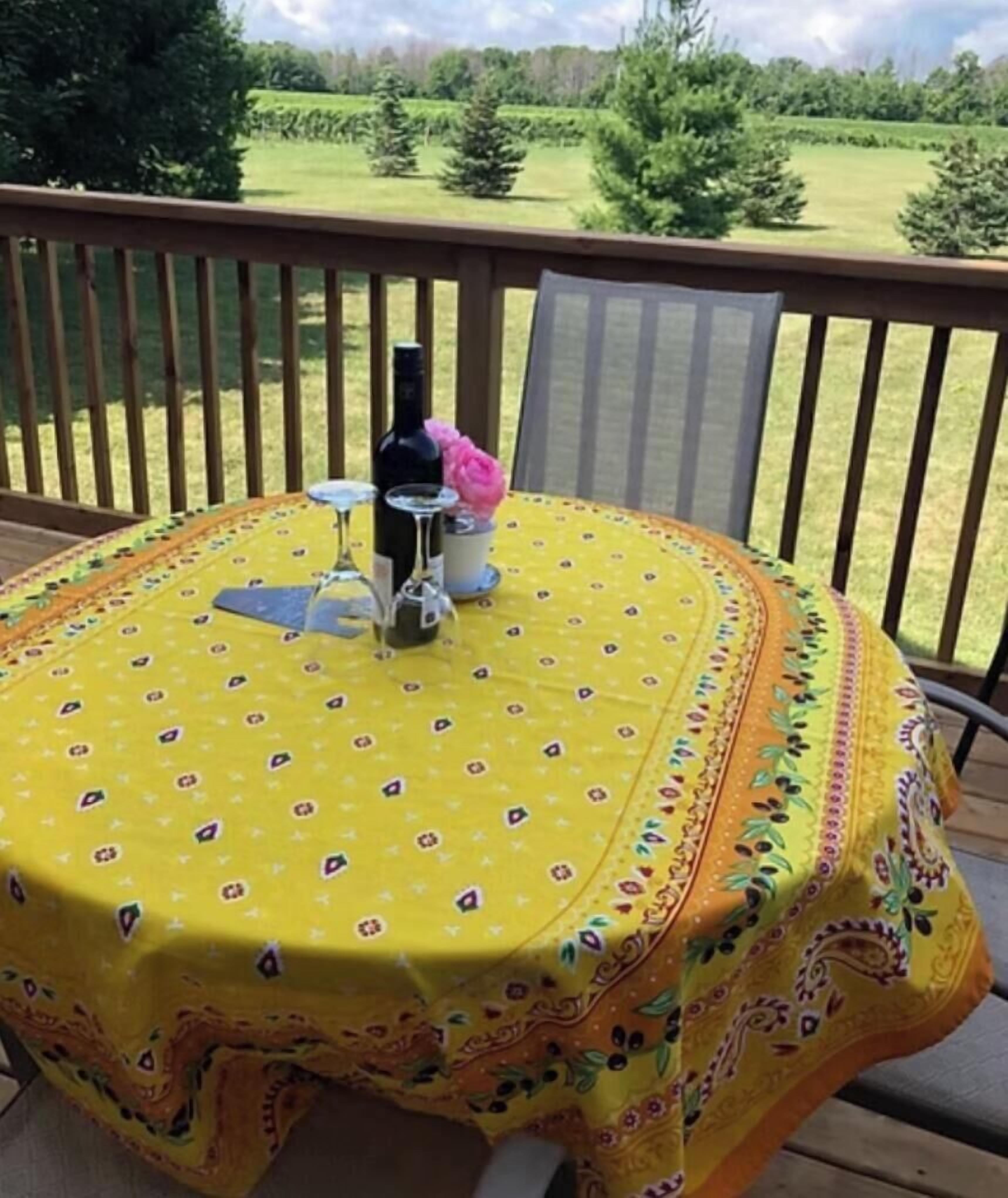 A round table with a yellow tablecloth, a bottle, and a glass on it, set on a wooden deck with a view of a green landscape.
