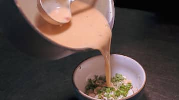 Een hand giet een romige vloeistof uit een pot in een kom met gehakte groenten.