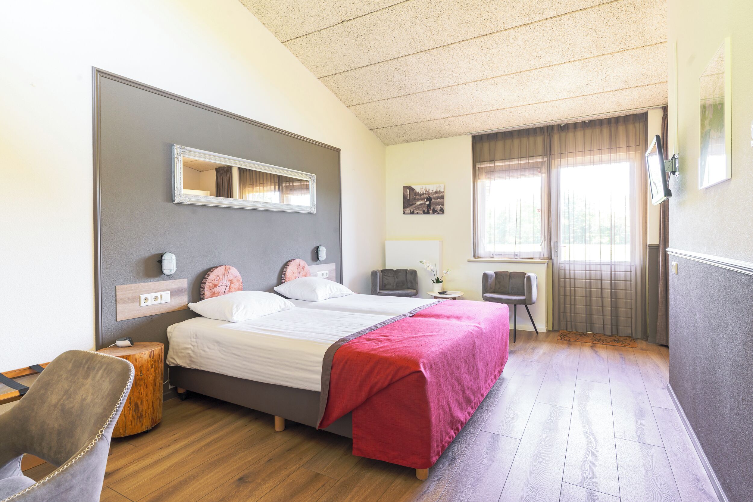 A hotel room with a bed, bedside tables, a chair, a desk, a mirror, and a window with curtains.
