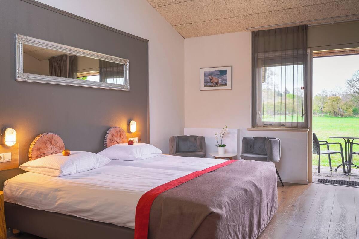 A hotel room with a large bed, a mirror, a small table with chairs, a window with a view of the outdoors, and a picture on the wall.