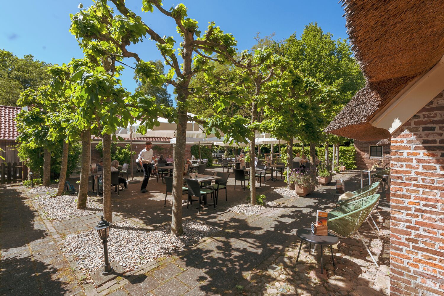 An outdoor patio area with tables and chairs, surrounded by trees and a brick building.