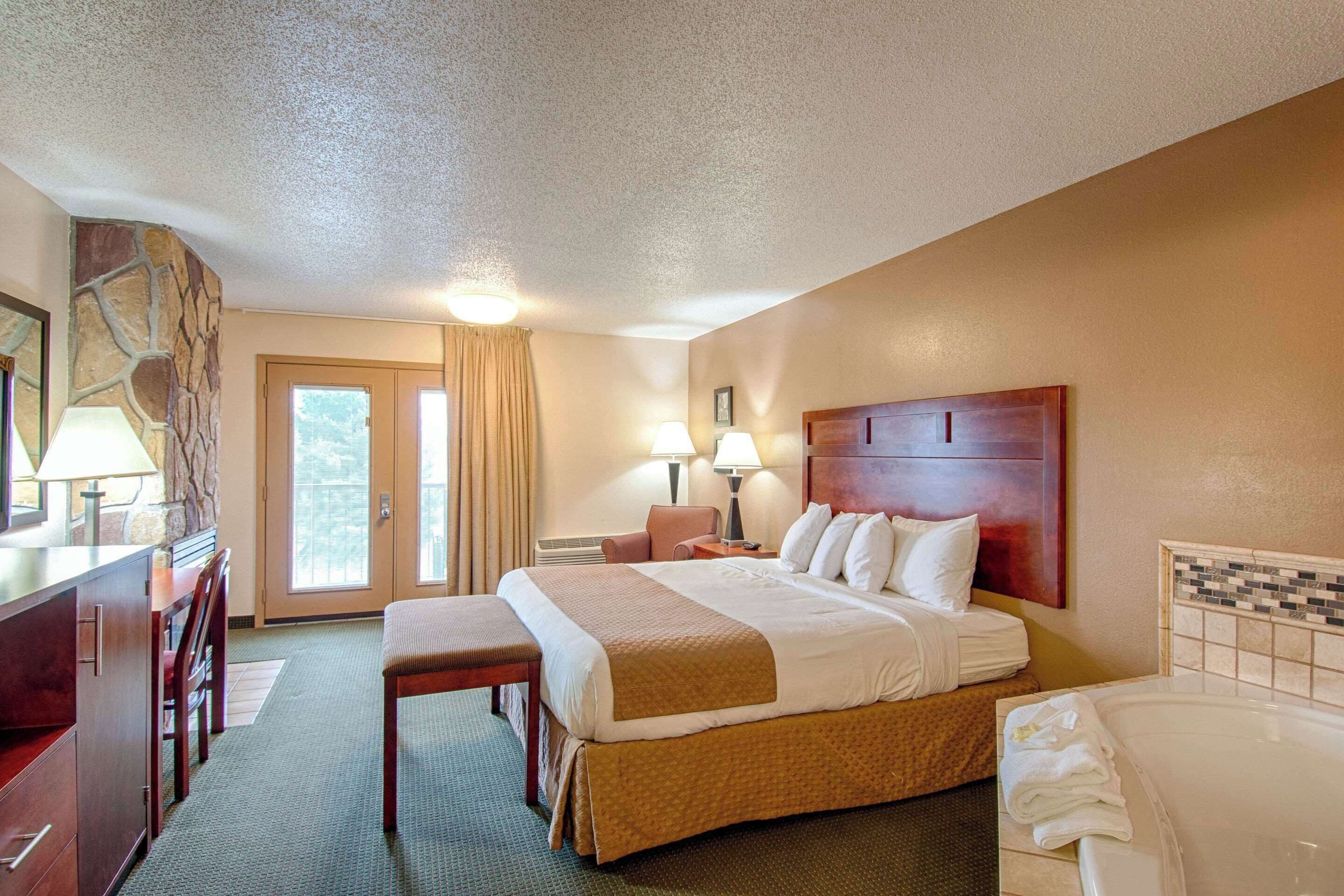 A hotel room with a large bed, a desk with a lamp, a chair, a bench, a bathroom with a bathtub, and a view of the outdoors through the door.