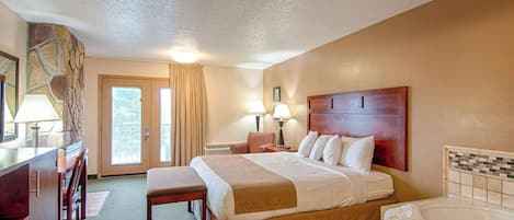 Habitación de hotel con una cama grande, un escritorio con lámpara, una silla, un banco, un baño con bañera y vistas al exterior desde la puerta.