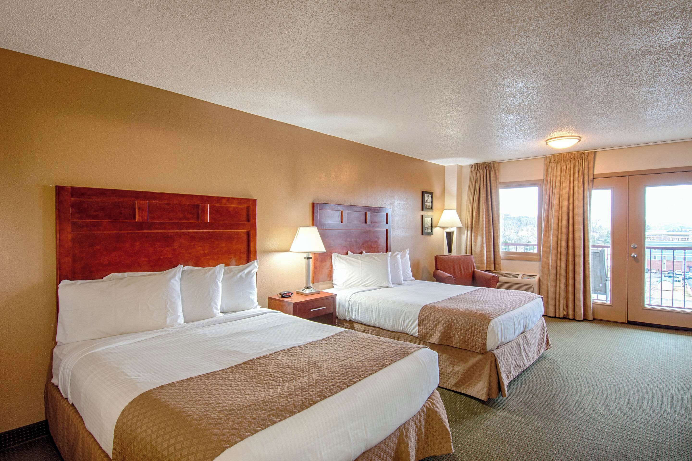 A hotel room with two beds, a wooden headboard, a nightstand with a lamp, and a view of the outdoors through a sliding door.