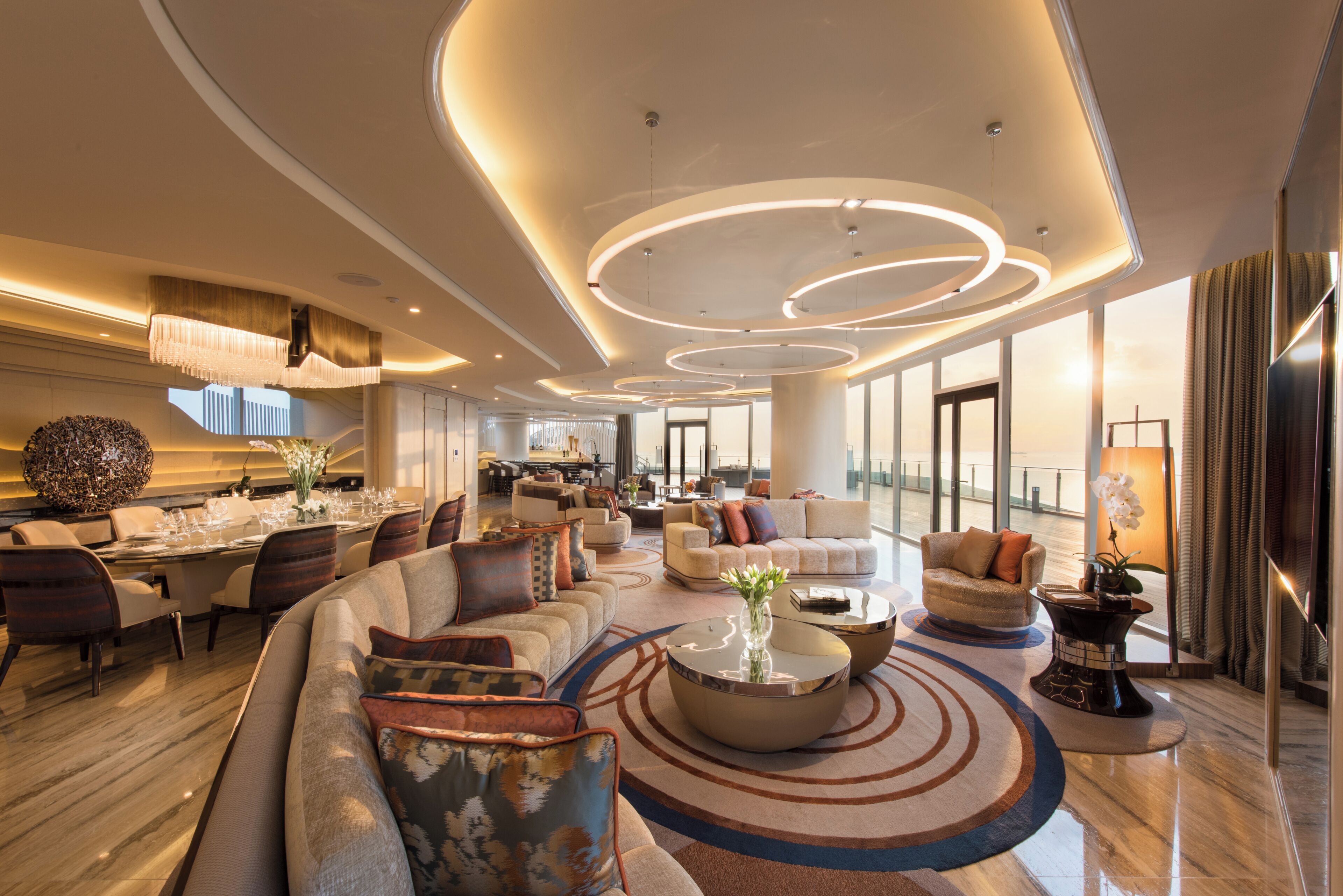 A spacious hotel lobby with a modern design, featuring a large seating area, a dining setup, and a view of the cityscape through the windows.