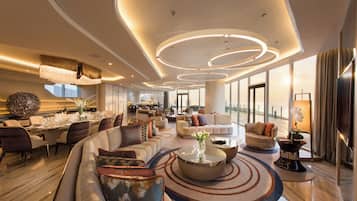 A spacious hotel lobby with a modern design, featuring a large seating area, a dining setup, and a view of the cityscape through the windows.