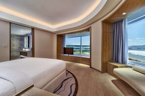 A modern hotel room with a large bed, a seating area, a TV, and a window with a view of the sea.