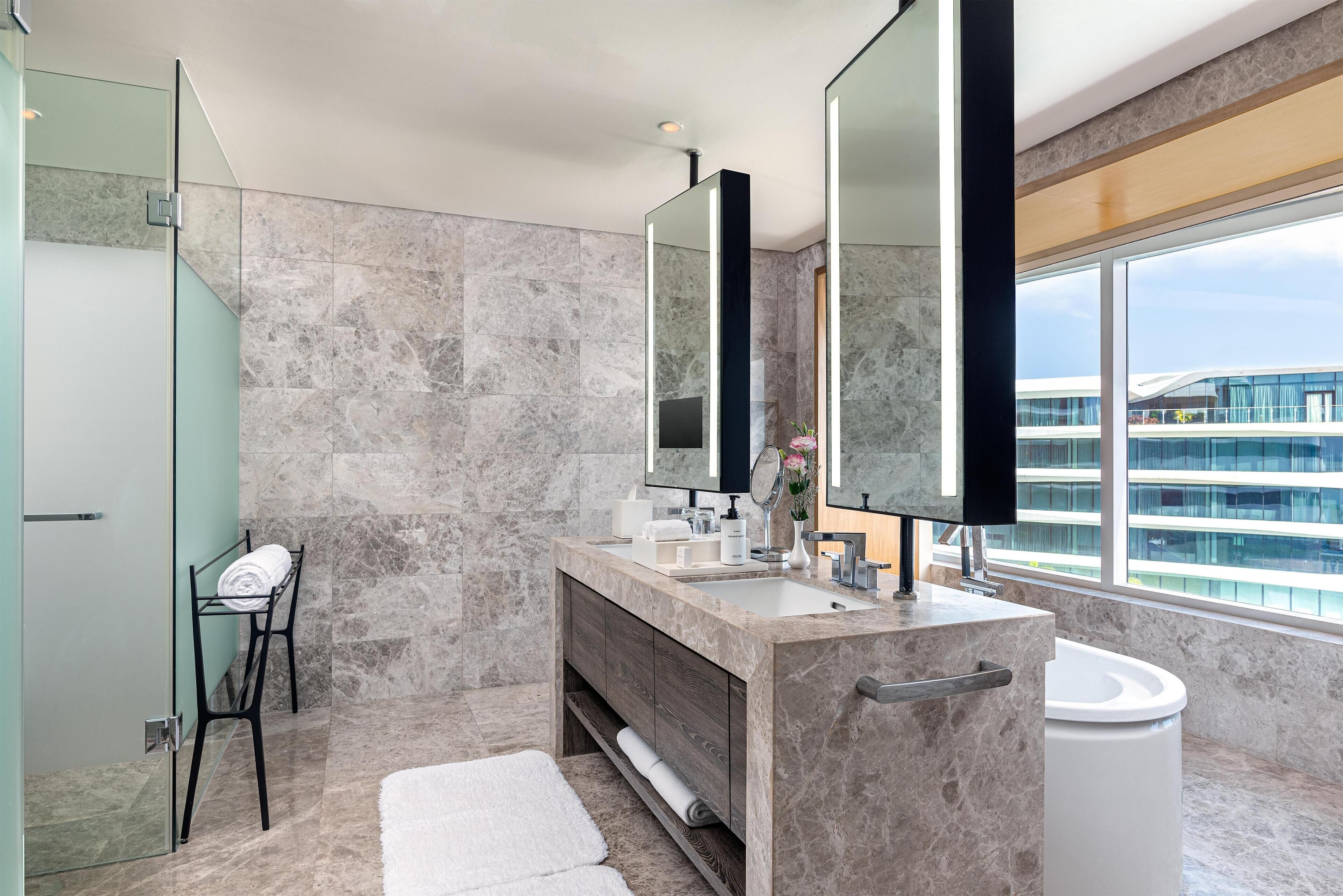 Un baño moderno con un lavamanos grande de mármol, cabina de ducha de vidrio y un ventanal con vistas a un edificio.