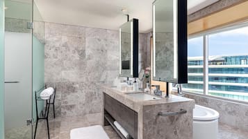 Un baño moderno con un lavamanos grande de mármol, cabina de ducha de vidrio y un ventanal con vistas a un edificio.