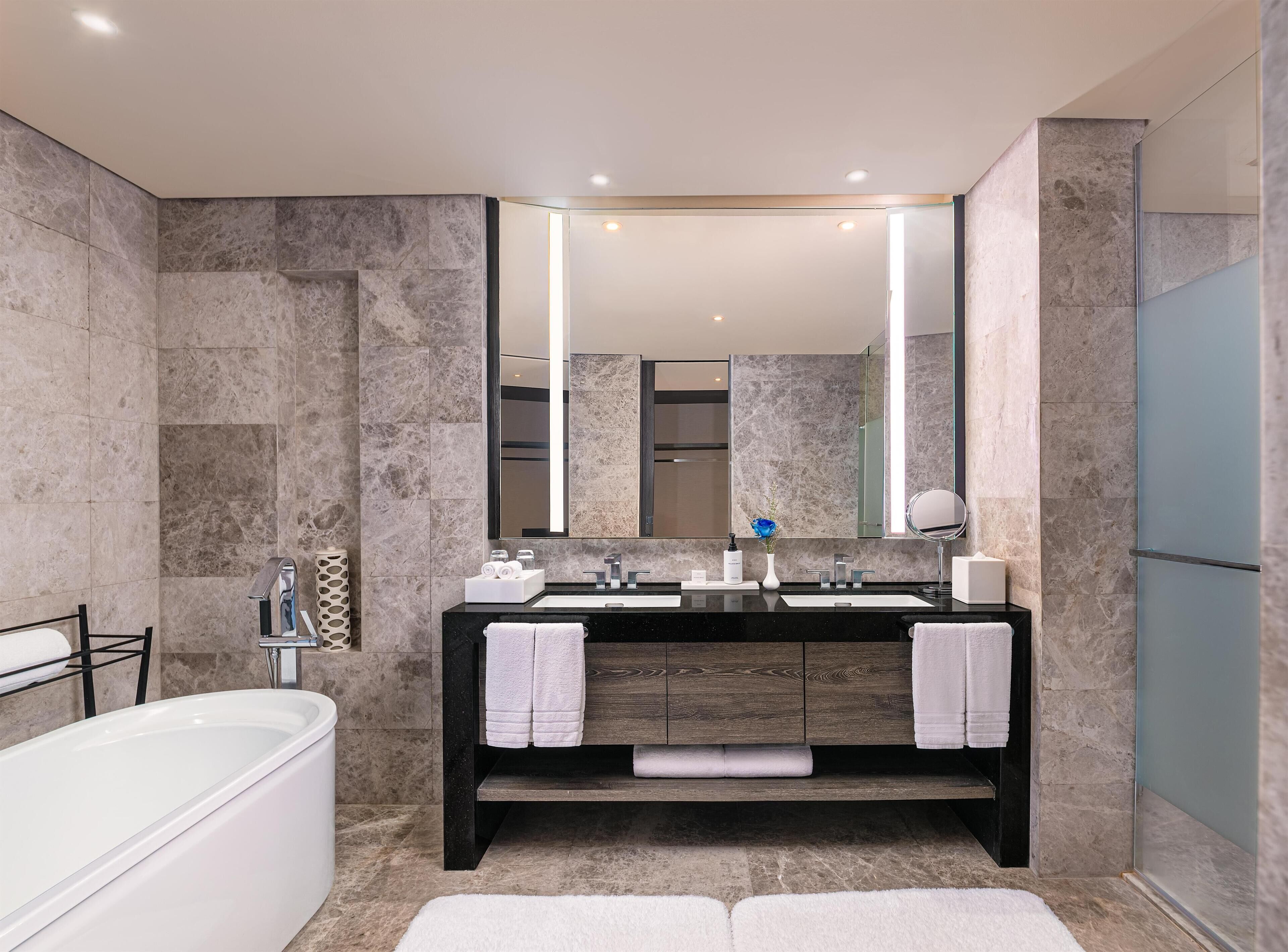 A modern bathroom with a large mirror, two sinks, a bathtub, and marble walls.