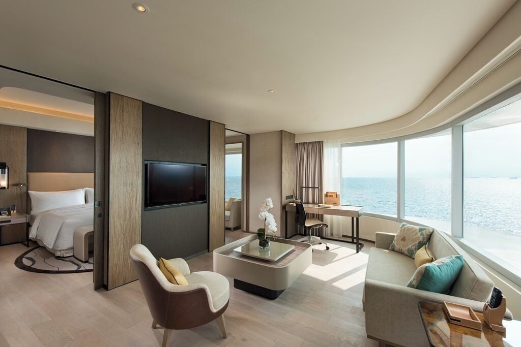 A modern hotel room with a large window, a flat-screen TV, a sofa, a coffee table, and a desk with a chair.