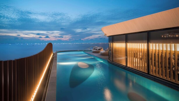 A modern infinity pool with a wooden deck, a glass-enclosed lounge area with warm lighting, and a view of the ocean at dusk. - Conrad Manila (Pasay)