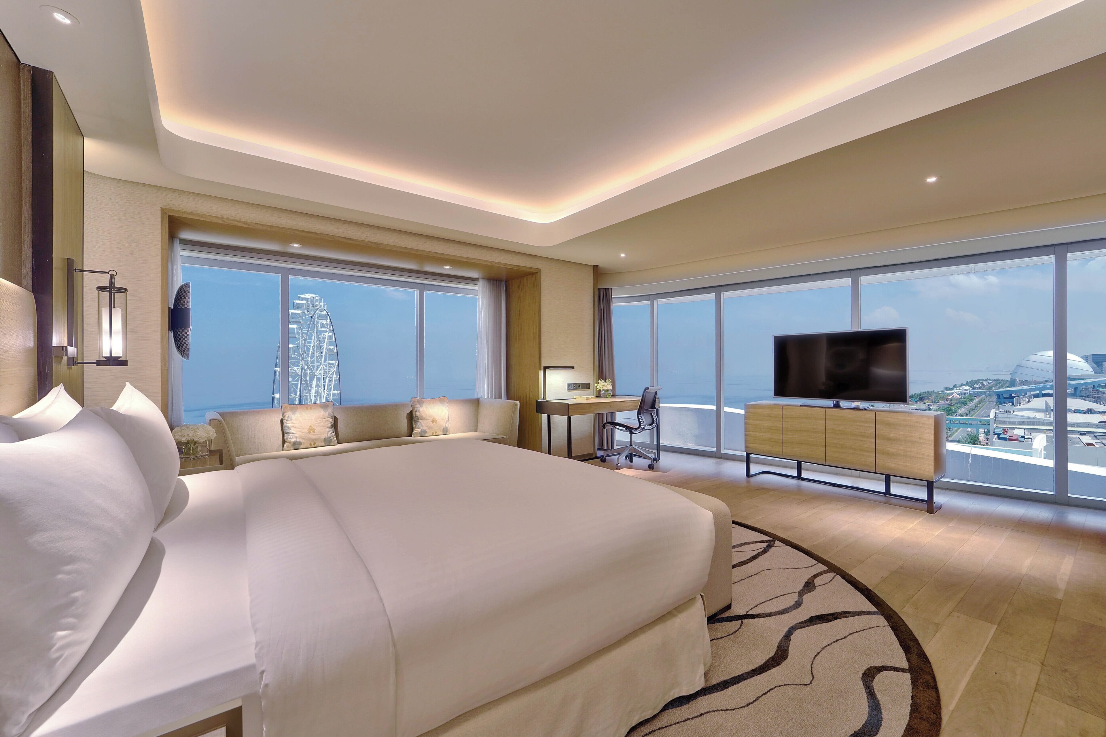 A modern hotel room with a large bed, a desk, a chair, a TV, and a view of a cityscape through large windows.