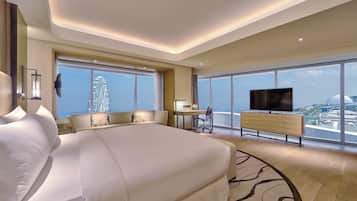 A modern hotel room with a large bed, a desk, a chair, a TV, and a view of a cityscape through large windows.