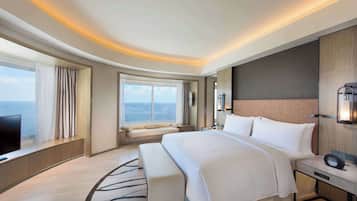 A modern hotel room with a large bed, a bench, a TV, and a view of the sea.