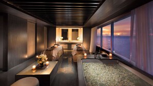 A modern spa room with a large bathtub, two massage tables, and a view of the sunset over the water. - Conrad Manila (Pasay)