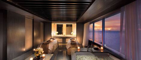 Une salle de spa moderne dotée d’une grande baignoire, de deux tables de massage et offrant une vue sur le coucher de soleil sur l’eau.
