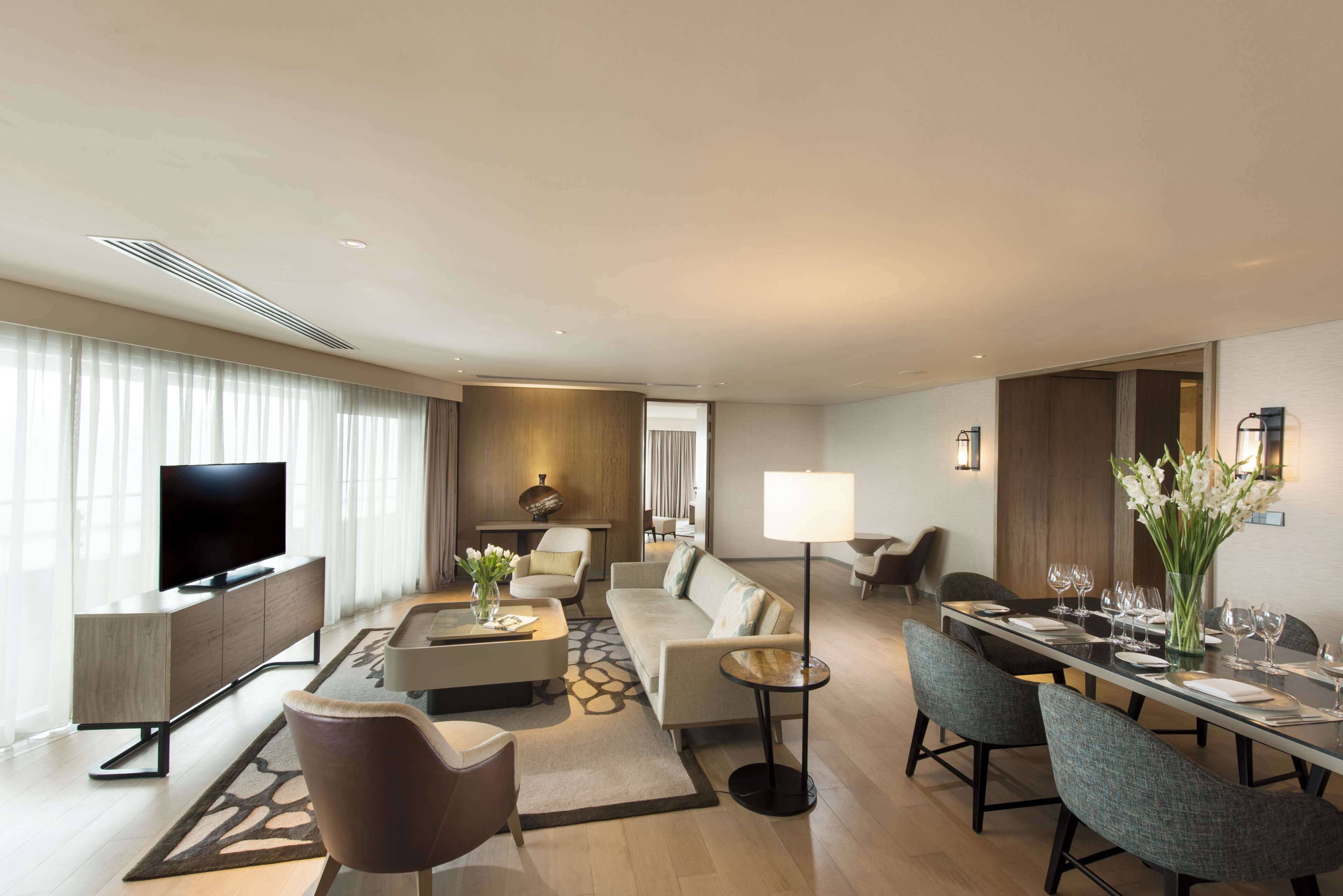 A modern hotel room with a flat-screen TV, a sofa, a dining table set for four, and a coffee table.