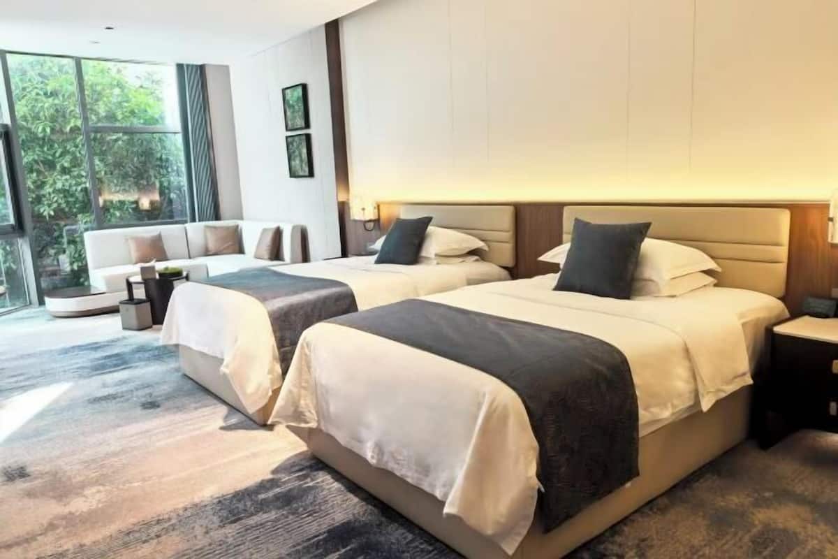 Une chambre d’hôtel avec deux lits, un canapé, une table de chevet et une grande fenêtre donnant sur un paysage verdoyant.