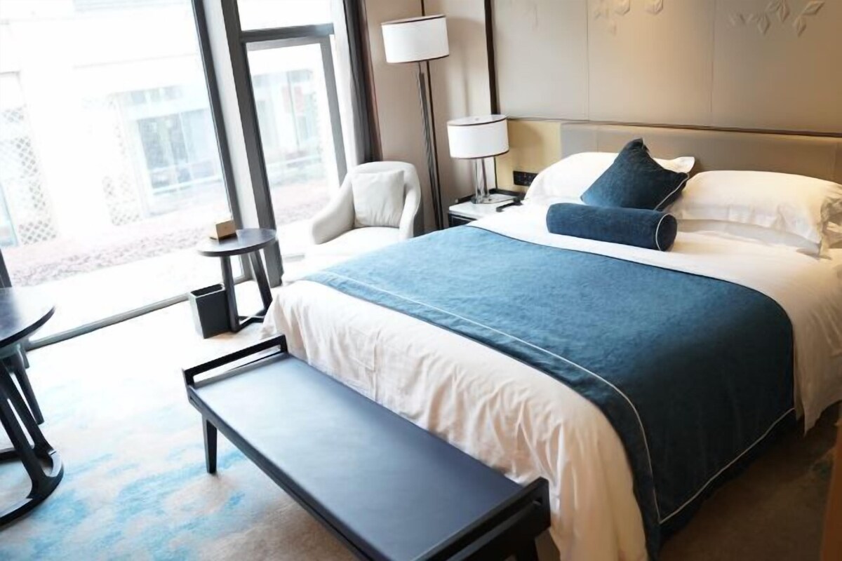 A hotel room with a large bed, a bench, a chair, a desk, and a lamp.