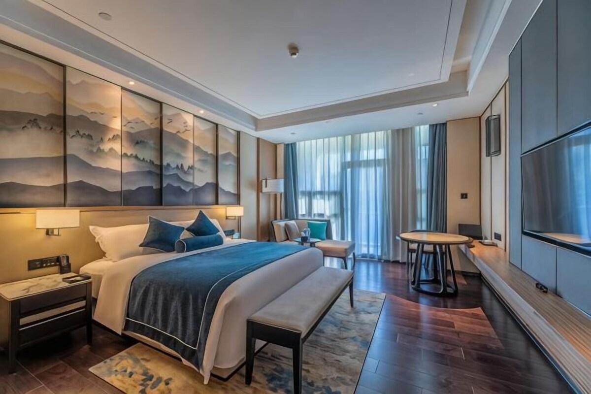 A modern hotel room with a large bed, bedside tables, a desk, a chair, a round table, and a TV.