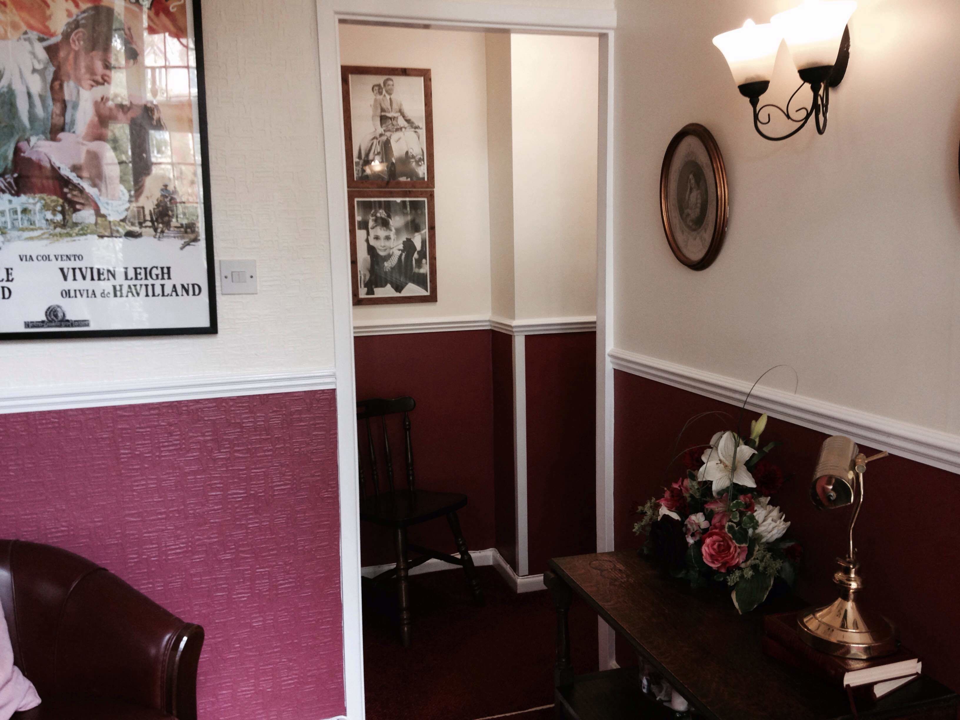 A room with a framed movie poster, a wall-mounted lamp, a desk with a vase of flowers, and a chair.