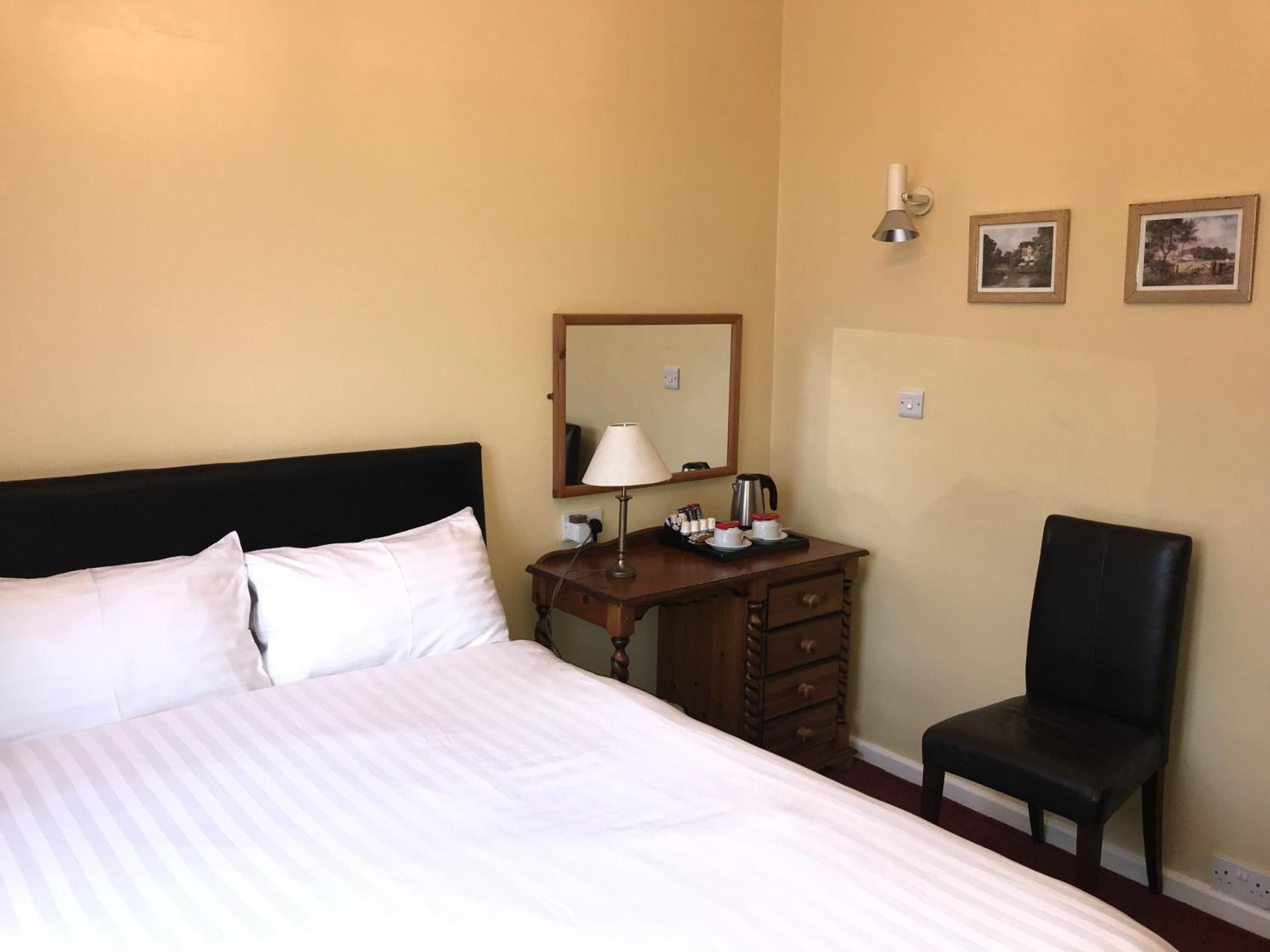 A hotel room with a bed, a wooden dresser, a chair, a lamp, and two framed pictures on the wall.