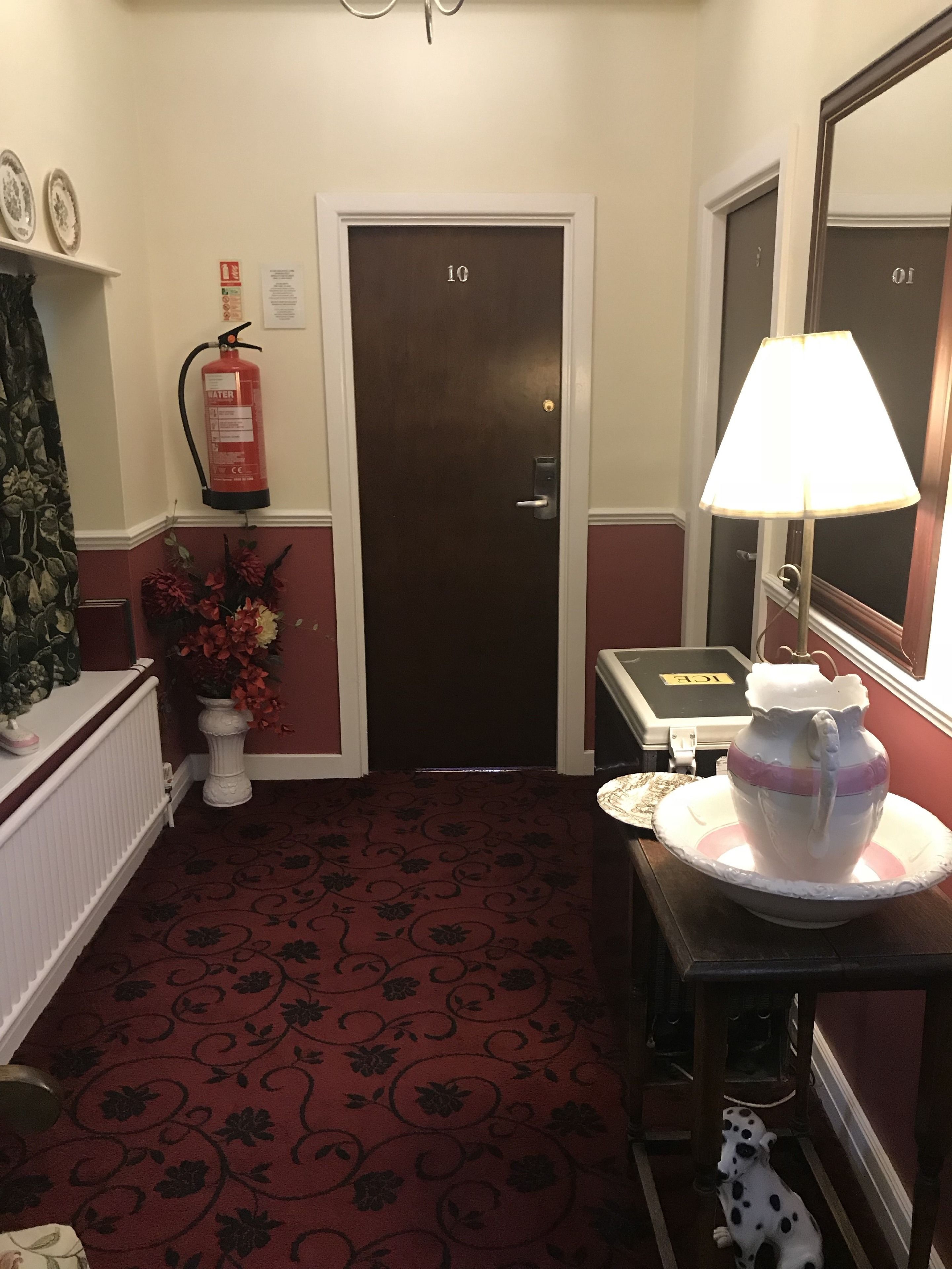 A hotel hallway with a dark brown door, a fire extinguisher, a lamp, and a small table with a decorative item.