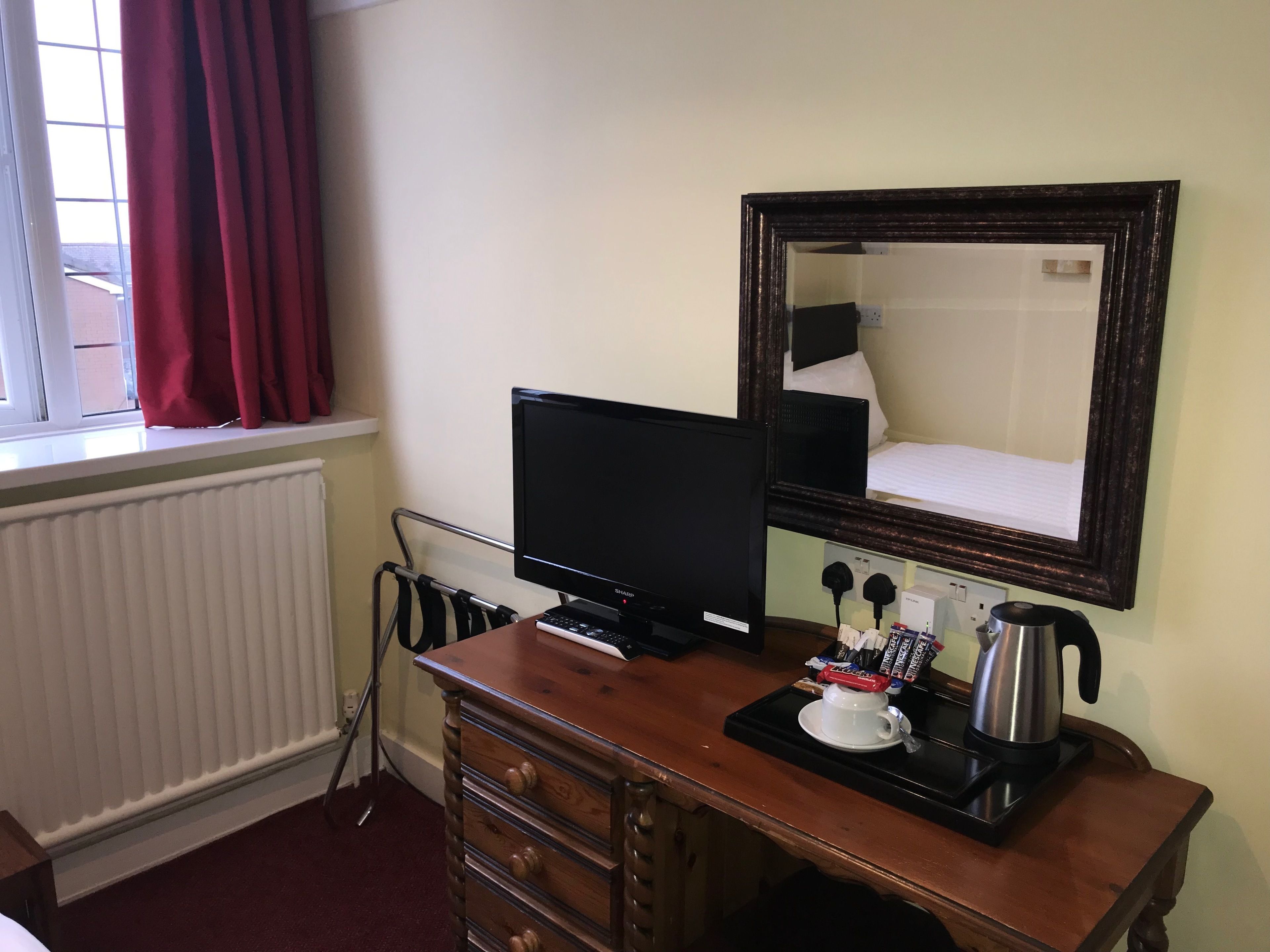 A hotel room with a wooden desk, a black monitor, a coffee maker, and a mirror on the wall.