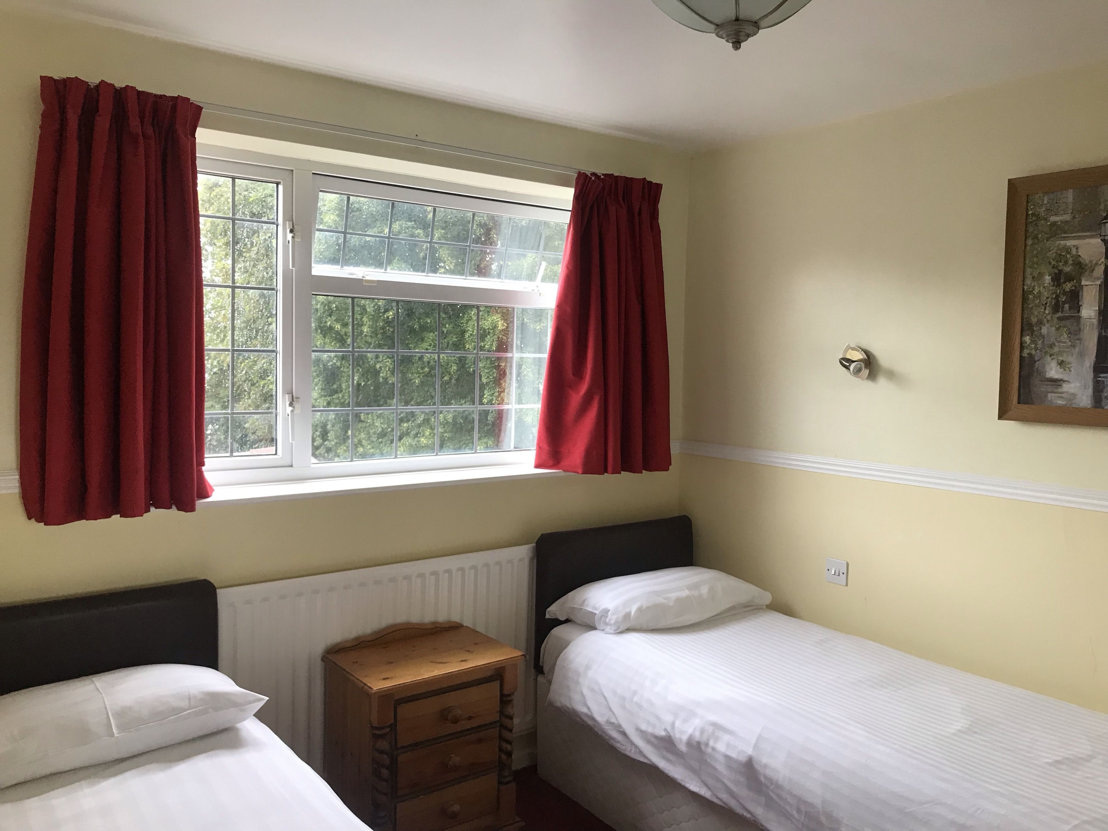 Un dormitorio con dos camas individuales, una mesita de noche de madera, un radiador y una ventana con cortinas rojas.