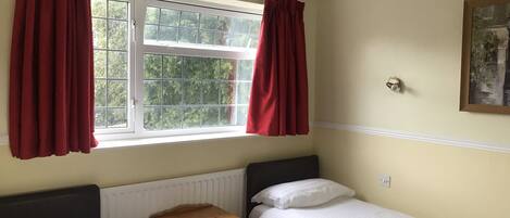 A bedroom with two single beds, a wooden nightstand, a radiator, and a window with red curtains.