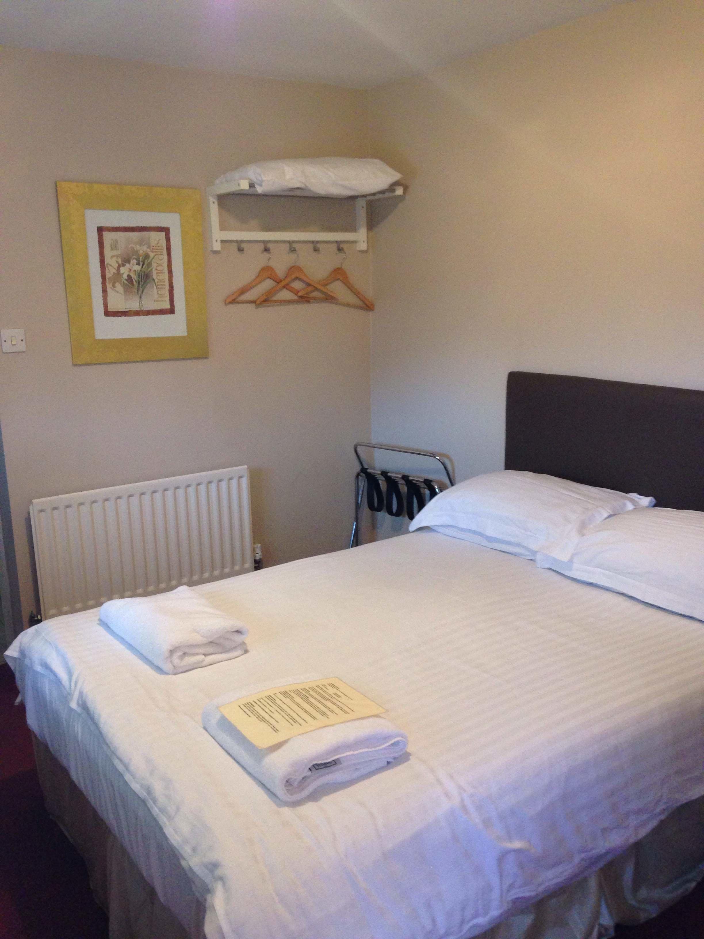 A neatly made bed with white linens, a folded towel, and a pillow. There is a radiator on the wall, a coat rack with hangers, and a framed picture above the bed.