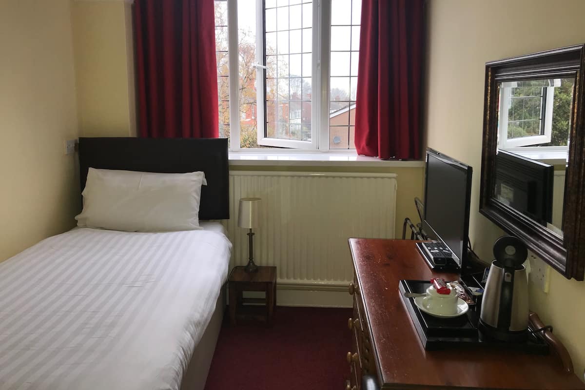 A hotel room with a bed, a small table with a kettle, a TV, and a window with red curtains.