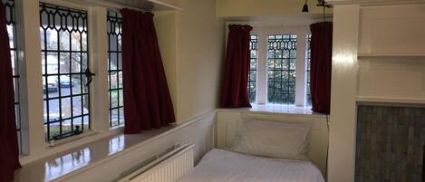 A bedroom with a bed, two windows with metal grilles, red curtains, a radiator, and a white ceiling with exposed beams.
