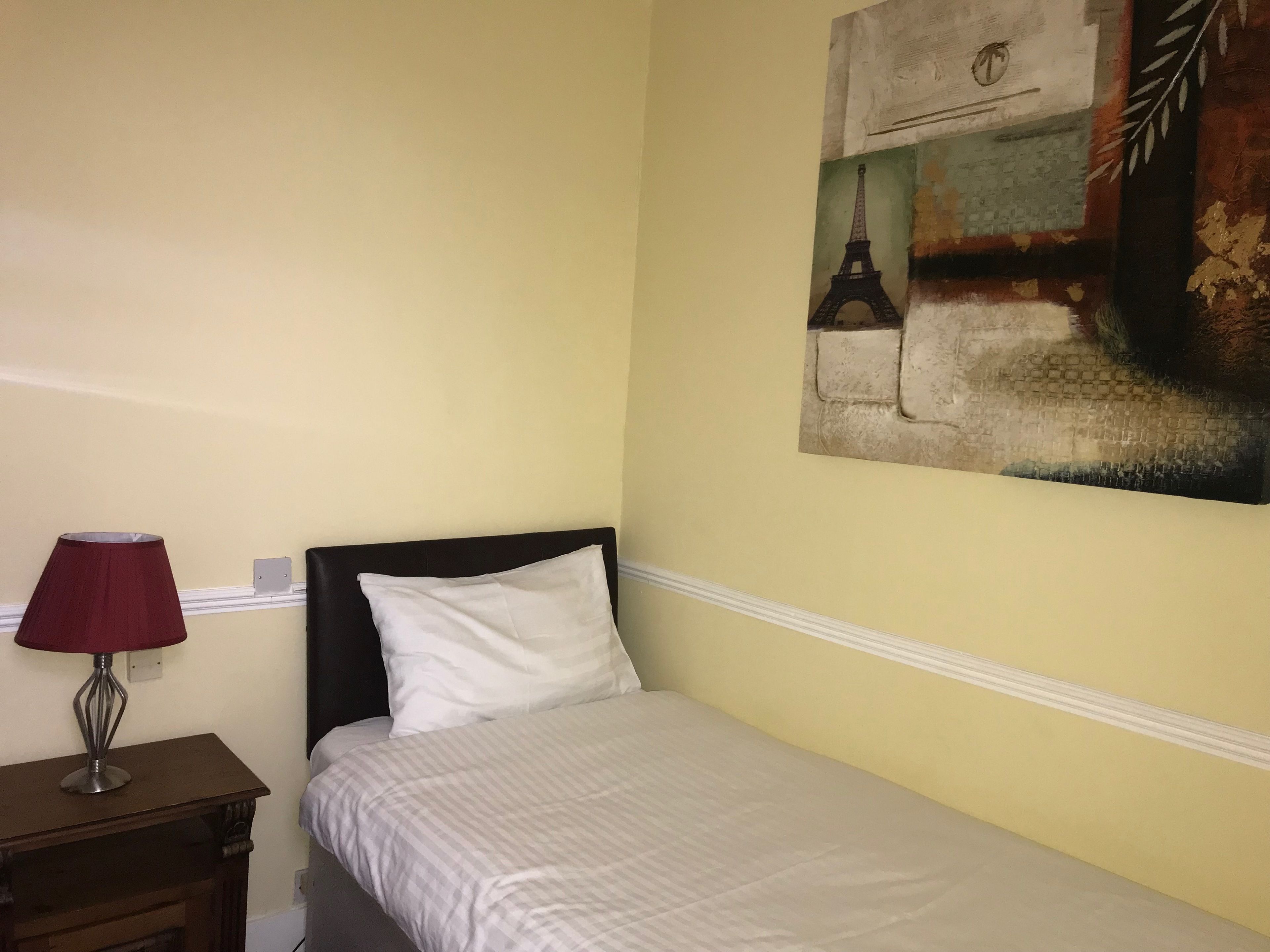 A bedroom with a bed, a nightstand, a lamp, and a wall painting of the Eiffel Tower.
