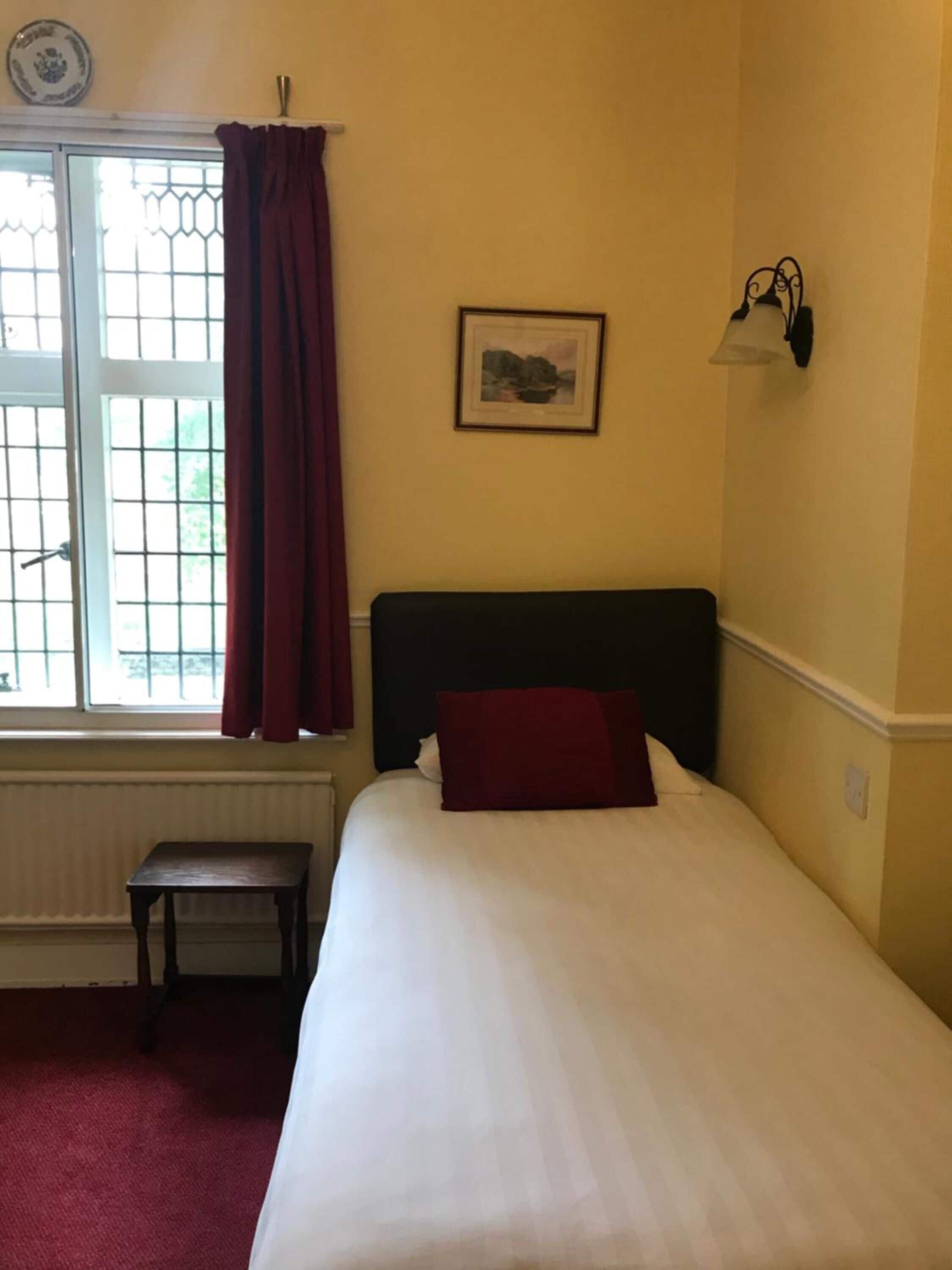 Une pièce comprenant un lit, une fenêtre avec des rideaux, une petite table et un luminaire fixé au mur.