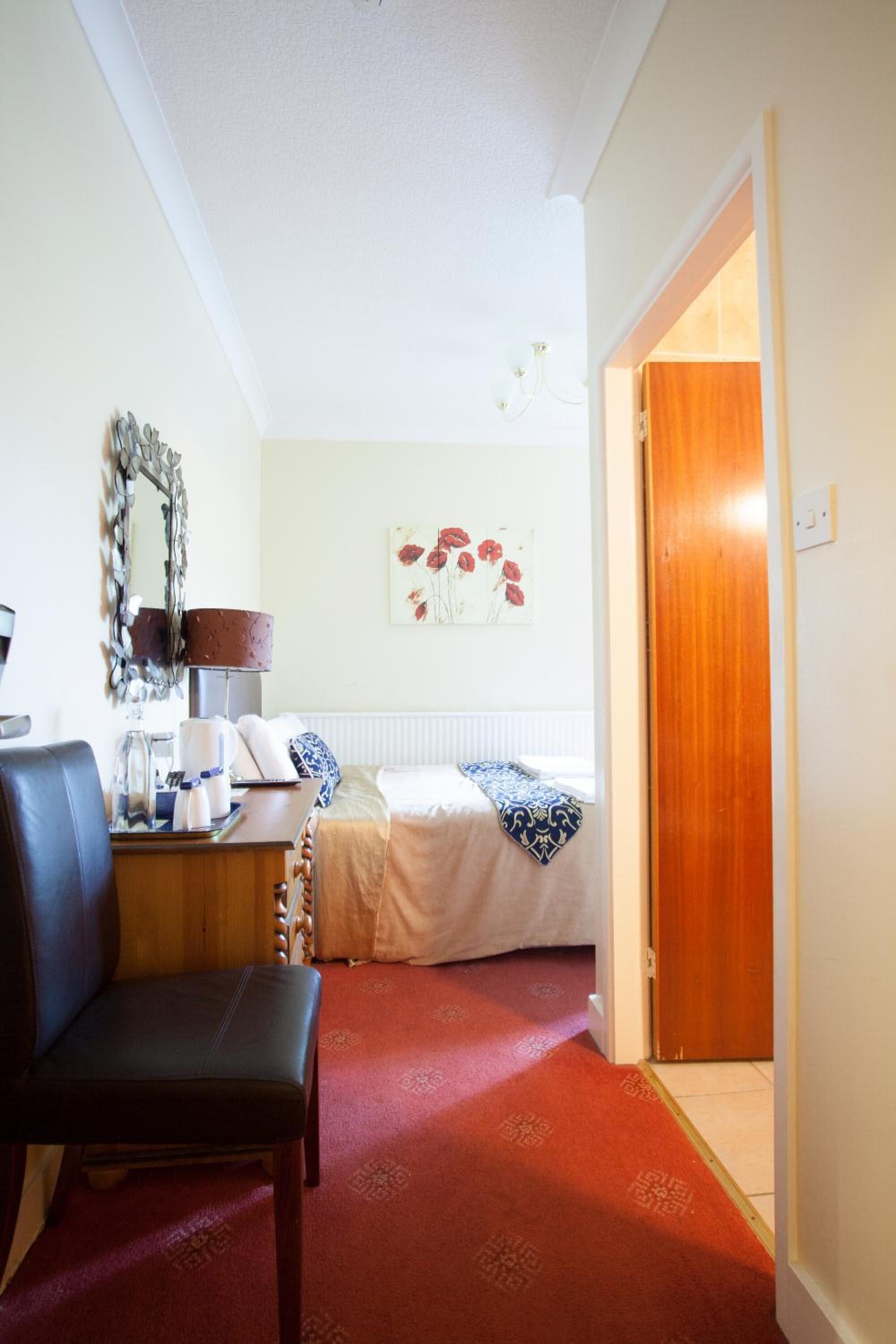 A hotel room with a bed, a desk with a lamp, a chair, a mirror, and a red carpet.