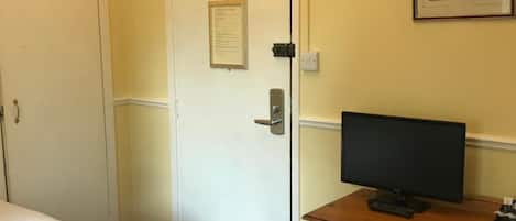 A hotel room with a bed, a desk with a computer, a trash bin, and a door with a notice.