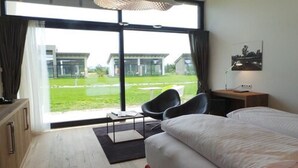 A modern hotel room with a large sliding glass door leading to a grassy outdoor area, a bed, a desk with a chair, and a small table with a lamp. - Hotel Diamond Country Club (Atzenbrugg)