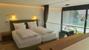 A double bed with white linens, two bedside tables, a wooden bench, a window with blinds, and a wall-mounted lamp.