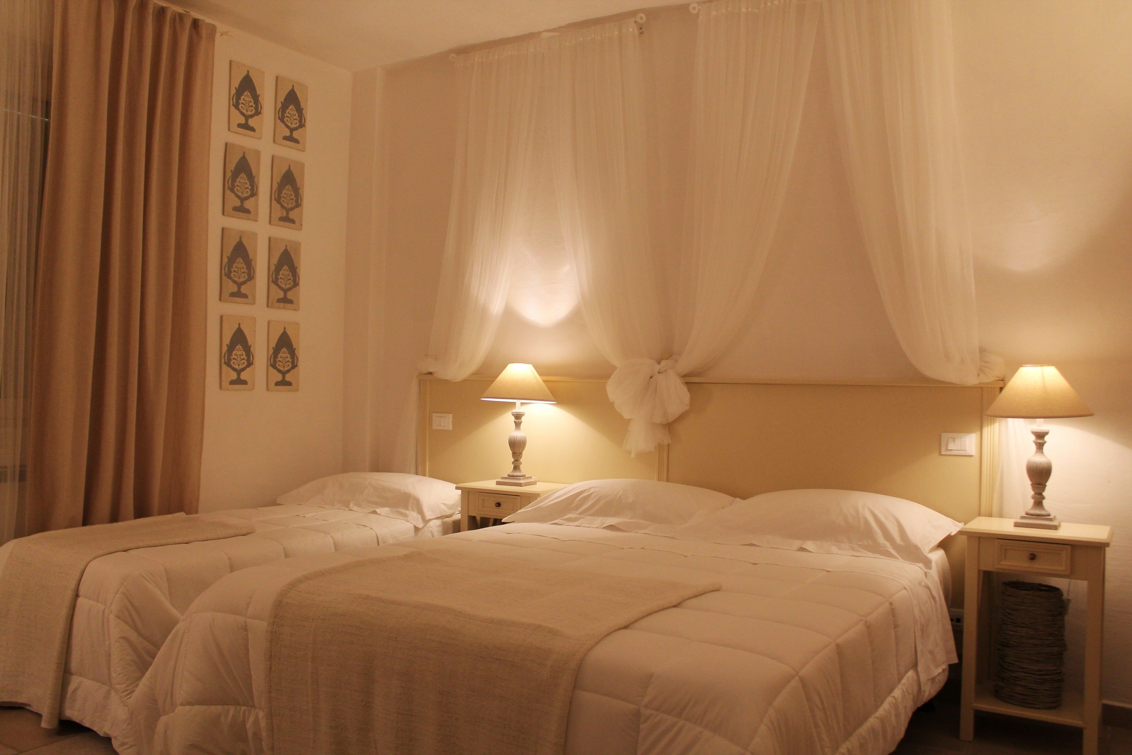 Habitación de hotel con dos camas, cada una con una mesita de noche y lámpara, y un ventanal con cortinas translúcidas.