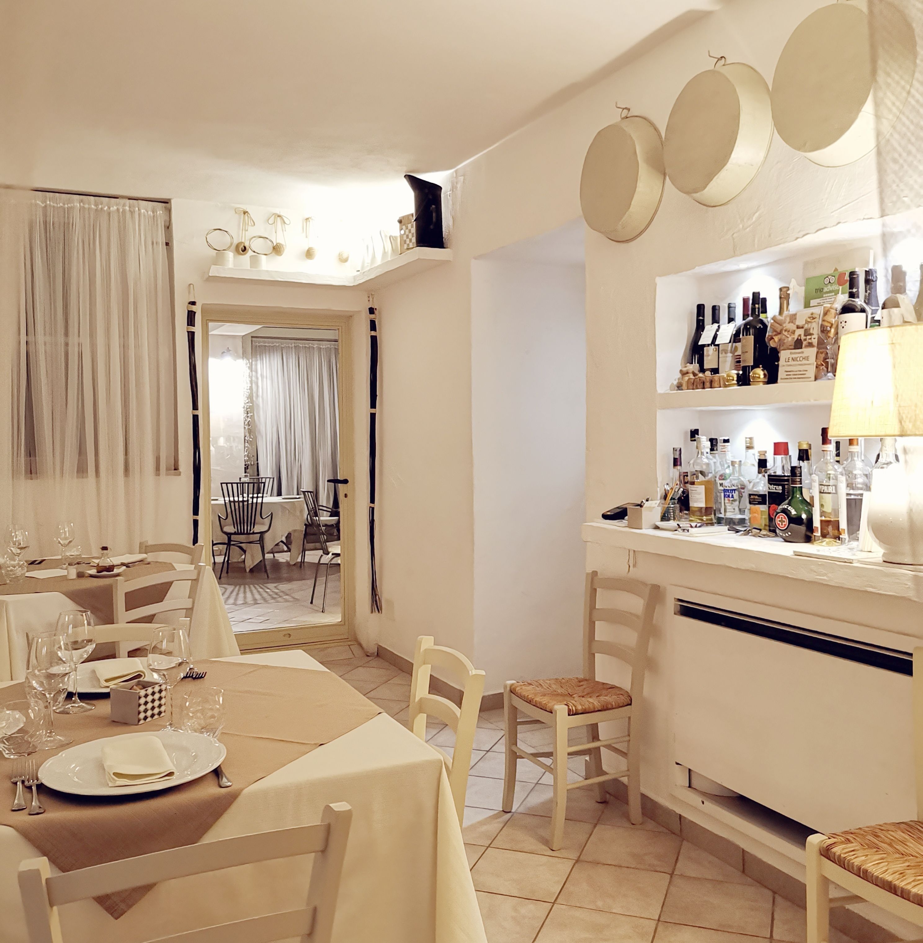 Angolo ristoro con tavolo apparecchiato per due, zona bar con diversi tipi di bottiglie, e vista su una sala da pranzo con tavolo e sedie.