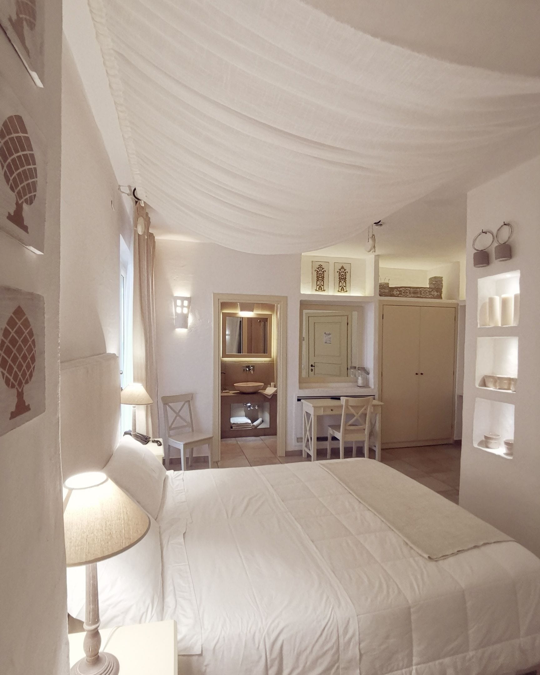 A bedroom with a bed, a desk, and a chair. There is a bathroom with a mirror and a sink. The room has white walls and a ceiling with draped fabric.