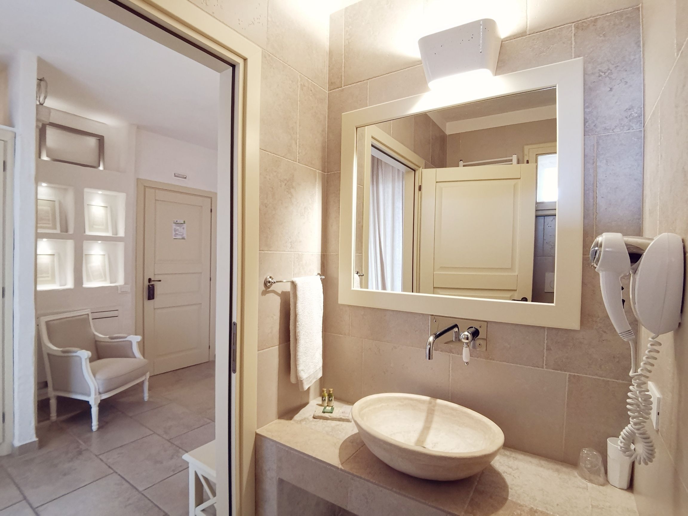 Une salle de bain avec un grand miroir, un lavabo et un sèche-cheveux. On aperçoit une chaise et une porte au fond.