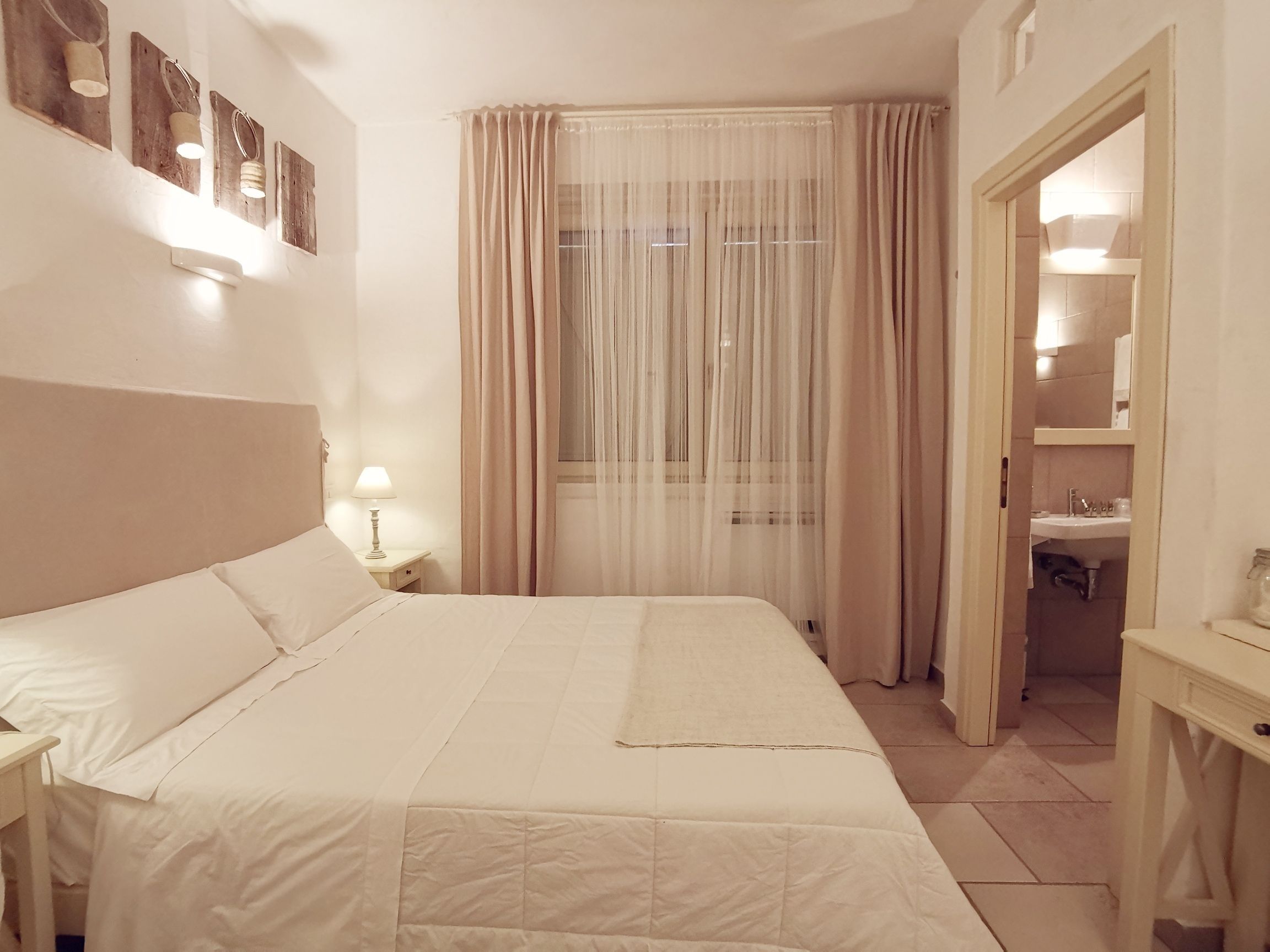Quarto de hotel com cama, mesa de cabeceira, luminária, janela com cortinas leves e banheiro visível através de uma porta aberta.