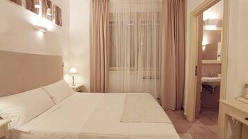 Quarto de hotel com cama, mesa de cabeceira, luminária, janela com cortinas leves e banheiro visível através de uma porta aberta.