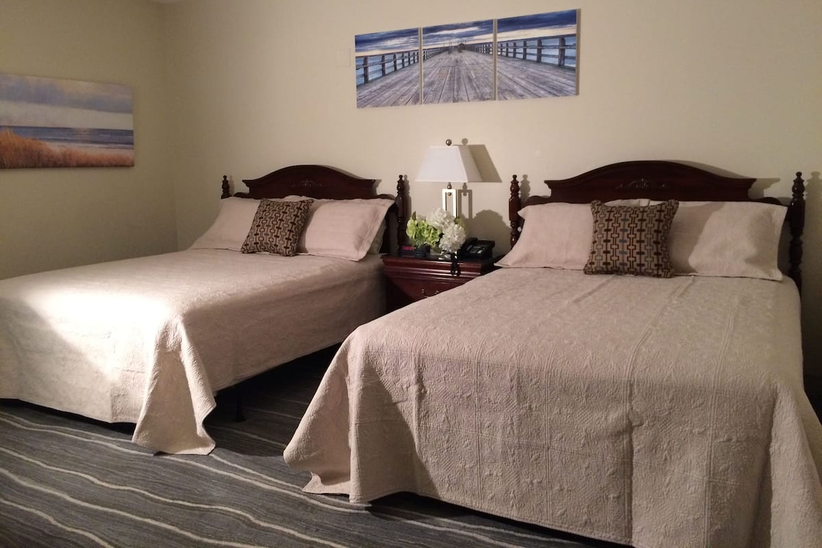 Une chambre d’hôtel avec deux lits, un tableau représentant une jetée, une lampe et une table de chevet avec un téléphone et des fleurs.